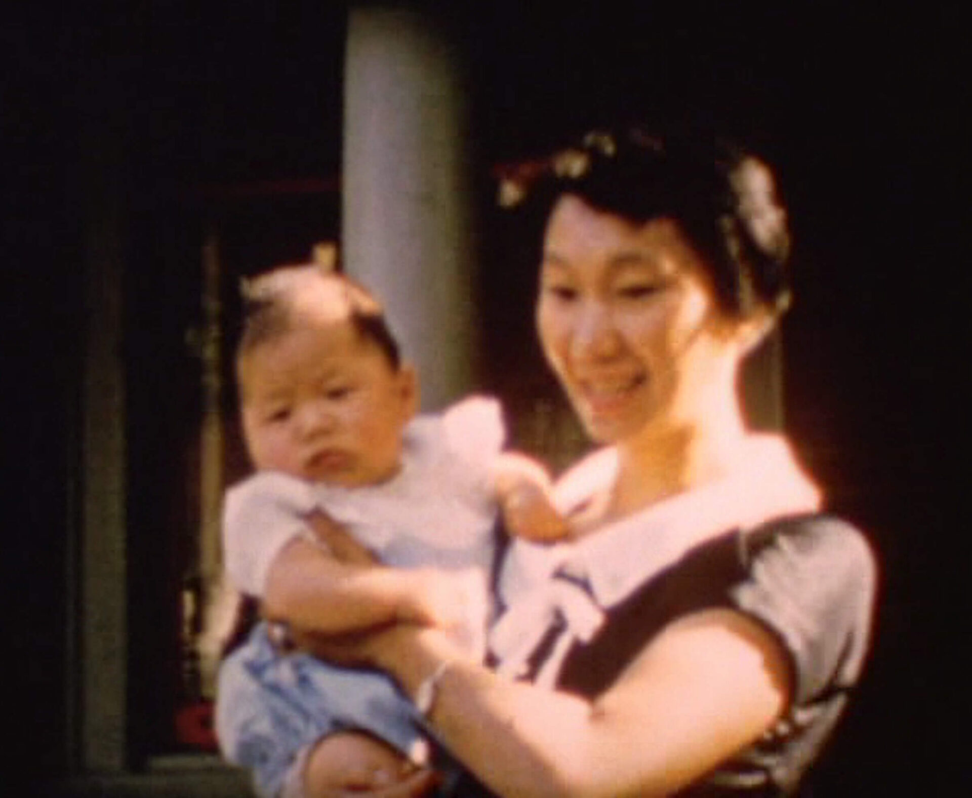 Still from The Dreaming House: A smiling woman holds a baby in her arms outside a house on a sunny day.