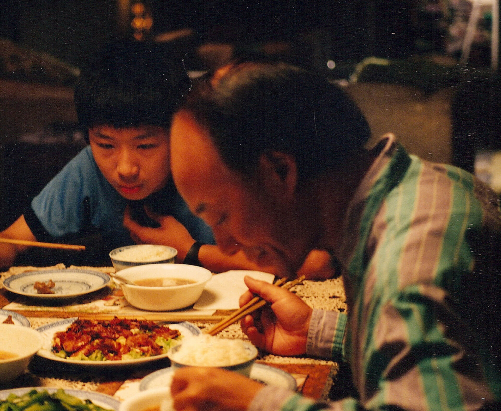 Still from Though Bananas: A man and a boy sit at a dinner table, both leaning slightly forward while eating with chopsticks, with several dishes of food on the table.